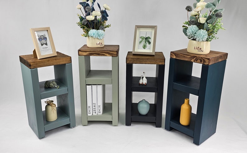 May include: Four small wooden side tables in varying colors: blue-gray, sage green, black, and navy. Each table has a dark brown top and a lower shelf. The tables are decorated with small objects and plants.