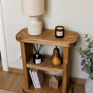 May include: A rustic, wooden side table with two shelves. The top shelf holds a beige lamp and a candle. The lower shelves display books, a wooden house, a pear-shaped ornament, and a diffuser. The table is stained a warm brown.