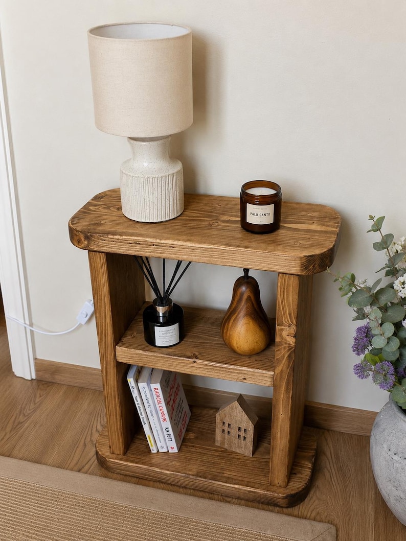May include: A rustic, wooden side table with two shelves. The top shelf holds a beige lamp and a candle. The lower shelves display books, a wooden house, a pear-shaped ornament, and a diffuser. The table is stained a warm brown.
