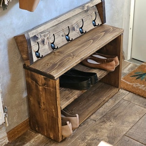 Op de afbeelding: Een houten schoenenrek met twee planken en een zitbank. Het rek heeft vijf haken aan de achterkant om jassen of tassen aan op te hangen. Het rek is in een donkerbruine kleur gebeitst.