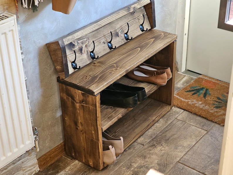 Op de afbeelding: Een houten schoenenrek met twee planken en een zitbank. Het rek heeft vijf haken aan de achterkant om jassen of tassen aan op te hangen. Het rek is in een donkerbruine kleur gebeitst.
