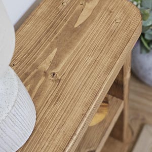 May include: A small, wooden, two-tiered shelf with a warm, brown stain. The top shelf has rounded corners and visible wood grain. The shelf is designed for decorative or storage purposes. The dimensions are approximately 12 inches long, 8 inches wide, and 10 inches tall.