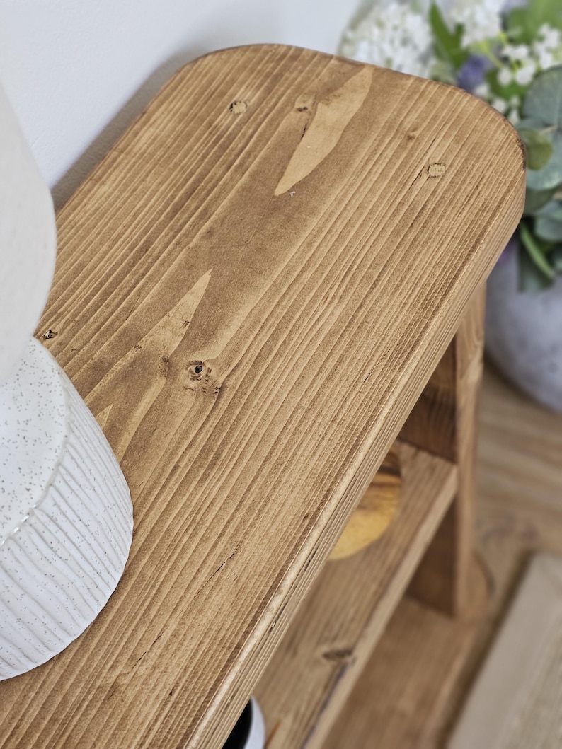 May include: A small, wooden, two-tiered shelf with a warm, brown stain. The top shelf has rounded corners and visible wood grain. The shelf is designed for decorative or storage purposes. The dimensions are approximately 12 inches long, 8 inches wide, and 10 inches tall.