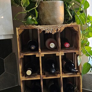 May include: A wooden wine rack with nine compartments, holding several wine bottles. The rack is made of light brown wood and has a rustic appearance. A potted plant sits on top of the rack, adding a touch of greenery.