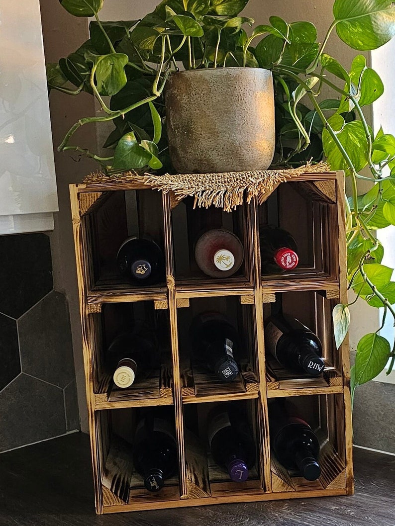 May include: A wooden wine rack with nine compartments, holding several wine bottles. The rack is made of light brown wood and has a rustic appearance. A potted plant sits on top of the rack, adding a touch of greenery.