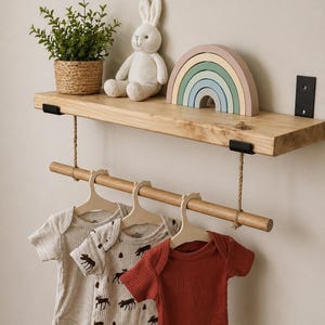 May include: A wooden shelf displays a small potted plant, a stuffed bunny, and a wooden rainbow. Below, three baby onesies hang on a wooden rod. One is rust-coloured, and two are beige with a moose pattern.