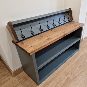 May include: A grey wooden shoe rack with a shelf and a top shelf with hooks. The top shelf is made of wood and has a dark stain. The hooks are black metal. The shoe rack is sitting on a wooden floor.