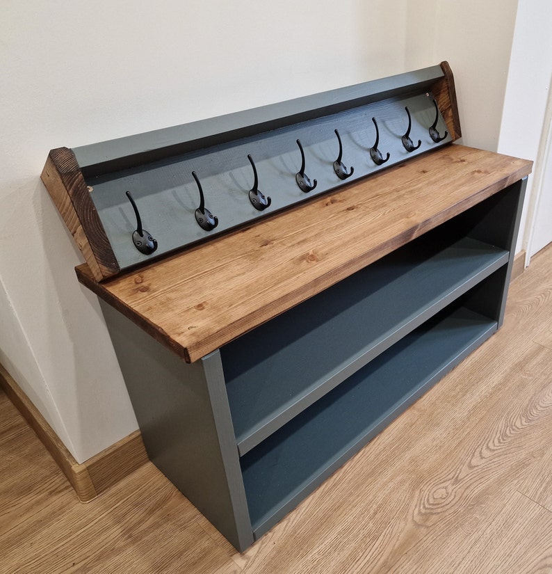 May include: A gray wooden shoe rack with a shelf and a top shelf with hooks. The top shelf is made of wood and has a dark stain. The hooks are black metal. The shoe rack is sitting on a wooden floor.