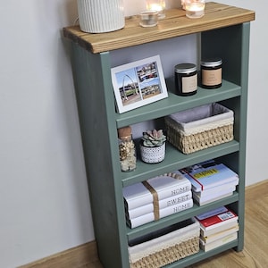 A wooden painted bookshelf with candles, framed photos, two baskets, decorative jars, and stacked books  placed on a wooden floor against a white wall.
