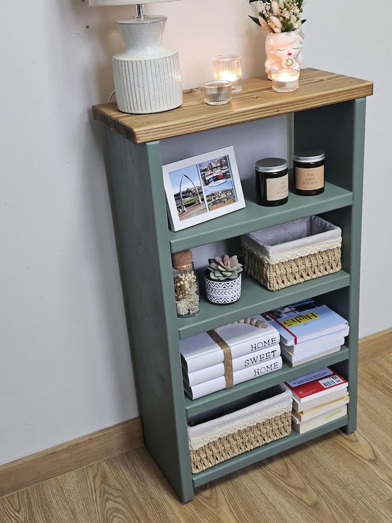 A wooden painted bookshelf with candles, framed photos, two baskets, decorative jars, and stacked books  placed on a wooden floor against a white wall.