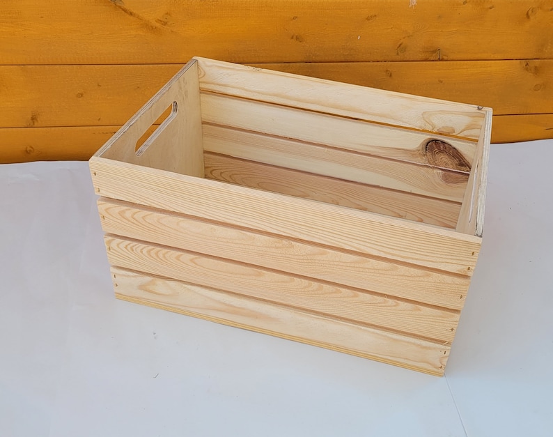 Puede incluir: Una caja de madera natural con forma rectangular y un asa en el lateral. La caja tiene un acabado de madera clara y est&aacute; hecha de madera sin tratar.