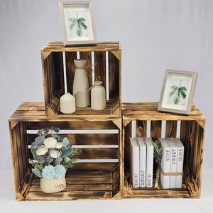 May include: Three wooden crates stacked on top of each other. The top crate has a framed print of a leaf, two white ceramic vases, and a smaller brown ceramic vase. The middle crate has a framed print of a leaf. The bottom crate has a bouquet of white flowers in a small white pot with the word "Life" on it. The bottom crate also has a stack of books with the words "Home Sweet Home" on them.
