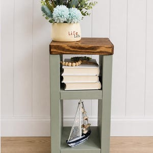 May include: A small, wooden side table with a dark brown top and sage green frame. The table holds a vase of flowers, books, and a decorative sailboat. The vase has the word "Life" on it.