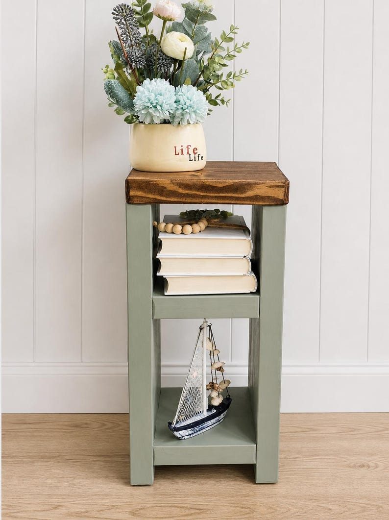May include: A small, wooden side table with a dark brown top and sage green frame. The table holds a vase of flowers, books, and a decorative sailboat. The vase has the word "Life" on it.