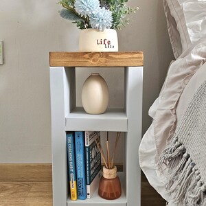 May include: A gray and wood bedside table with a vase of flowers and books. The table has two shelves and a wooden top. A vase with the word "Life" is on top. The books are titled "The Coaching Habit" and "Difficult Conversations".