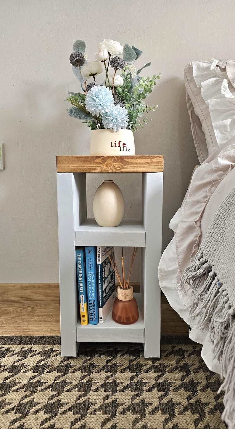 May include: A gray and wood bedside table with a vase of flowers and books. The table has two shelves and a wooden top. A vase with the word "Life" is on top. The books are titled "The Coaching Habit" and "Difficult Conversations".