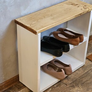 Op de afbeelding: Een witte houten schoenenrek met twee planken. De bovenste plank is van hout en heeft een natuurlijke afwerking. De onderste plank is wit en heeft een bruin kleed ervoor. Er staan vier paar schoenen op de planken, twee paar bruine ballerina's en twee paar zwarte ballerina's.