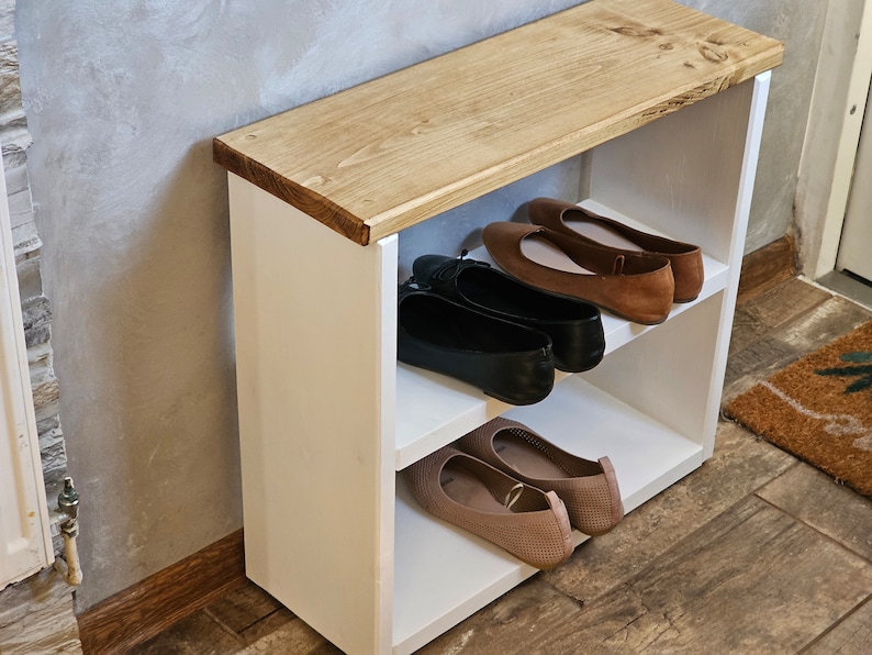 Op de afbeelding: Een witte houten schoenenrek met twee planken. De bovenste plank is van hout en heeft een natuurlijke afwerking. De onderste plank is wit en heeft een bruin kleed ervoor. Er staan vier paar schoenen op de planken, twee paar bruine ballerina's en twee paar zwarte ballerina's.