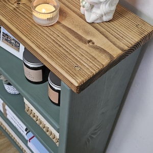 A wooden painted bookshelf with candles, framed photos, two baskets, decorative jars, and stacked books  placed on a wooden floor against a white wall.