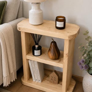 May include: A light-coloured wooden side table with two shelves. The top shelf holds a lamp with a beige shade and a candle. The lower shelves display books, a wooden pear, a diffuser, and a small wooden house.