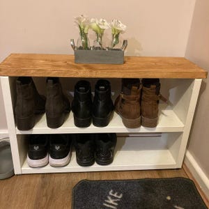 May include: A white wooden shoe rack with two shelves. The top shelf has a wooden top and is holding a vase with white flowers. The bottom shelf is holding a variety of shoes, including brown boots, black boots, and white sneakers.
