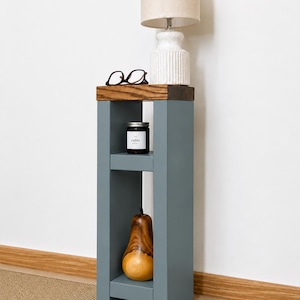 May include: A gray wooden side table with a lamp, glasses, a candle, and a wooden pear. The lamp has a beige lampshade and a white base. The candle jar has the word "calm" on it. The pear is brown and yellow.