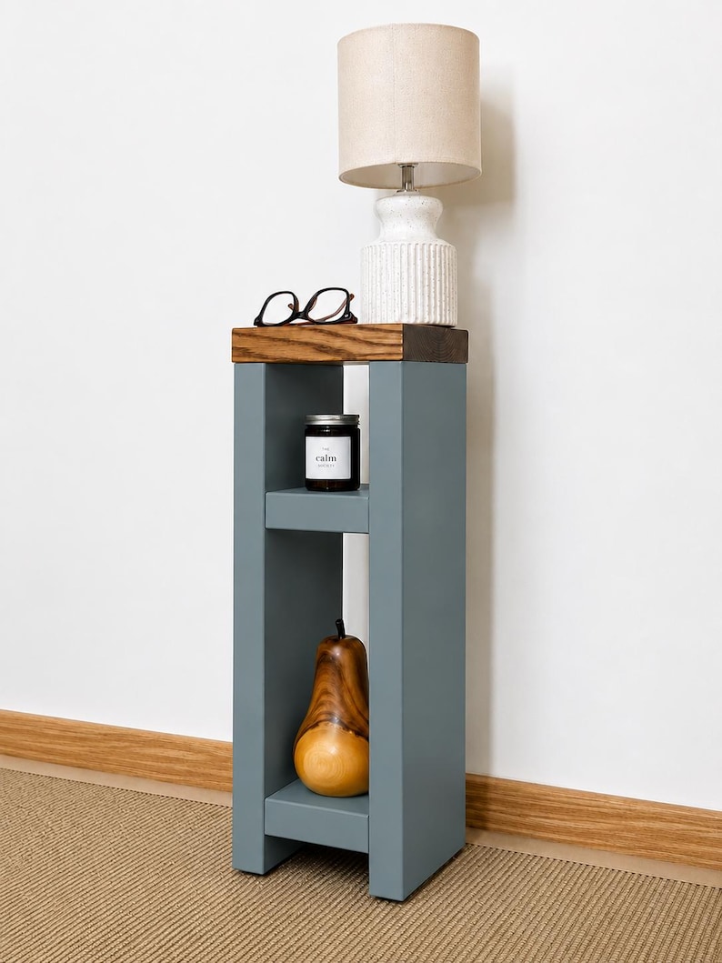 May include: A gray wooden side table with a lamp, glasses, a candle, and a wooden pear. The lamp has a beige lampshade and a white base. The candle jar has the word "calm" on it. The pear is brown and yellow.
