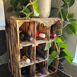 May include: A rustic, wooden wine rack with a charred finish, holding several wine bottles. The rack is made of square compartments and is topped with a potted plant. The wood grain is visible, and the overall aesthetic is natural and handcrafted.