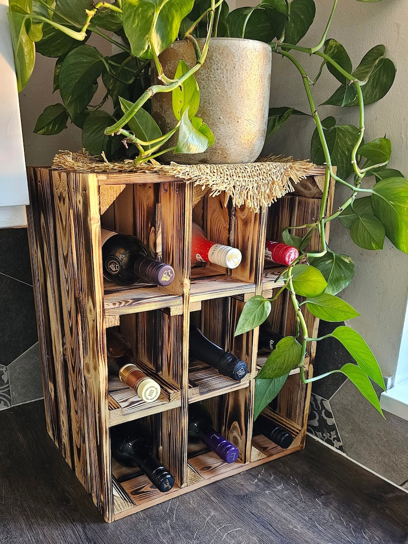 May include: A rustic, wooden wine rack with a charred finish, holding several wine bottles. The rack is made of square compartments and is topped with a potted plant. The wood grain is visible, and the overall aesthetic is natural and handcrafted.