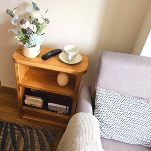 May include: A wooden two-tiered shelf unit with a white ceramic cup and saucer on the top shelf. The shelf is next to a gray armchair with a white and blue patterned cushion.