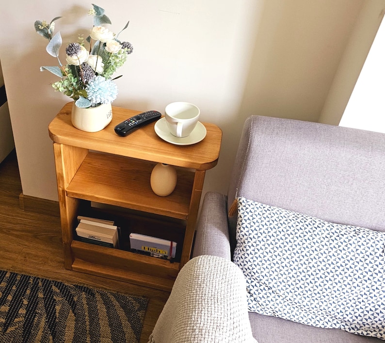 May include: A wooden two-tiered shelf unit with a white ceramic cup and saucer on the top shelf. The shelf is next to a gray armchair with a white and blue patterned cushion.