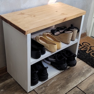 May include: White wooden shoe rack with two shelves. The top shelf is made of wood and has a natural finish. The bottom shelf is white and has a variety of shoes on it, including sneakers, flats, and sandals.