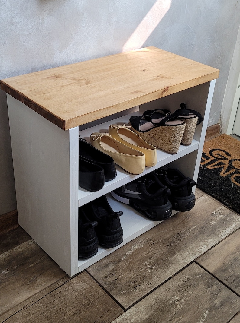 May include: White wooden shoe rack with two shelves. The top shelf is made of wood and has a natural finish. The bottom shelf is white and has a variety of shoes on it, including sneakers, flats, and sandals.