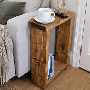 May include: A wooden side table with a warm, brown finish. The table has a rectangular top and a lower shelf, ideal for storing books. A white teacup and saucer sit on top. The table is next to a light grey sofa. The table is approximately 60 cm tall.