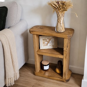 May include: A rustic, two-tiered wooden side table with a warm, honey-toned finish. The table holds a framed picture, a candle, and a decorative wooden pear. A clear glass vase with dried wheat stalks sits on top.