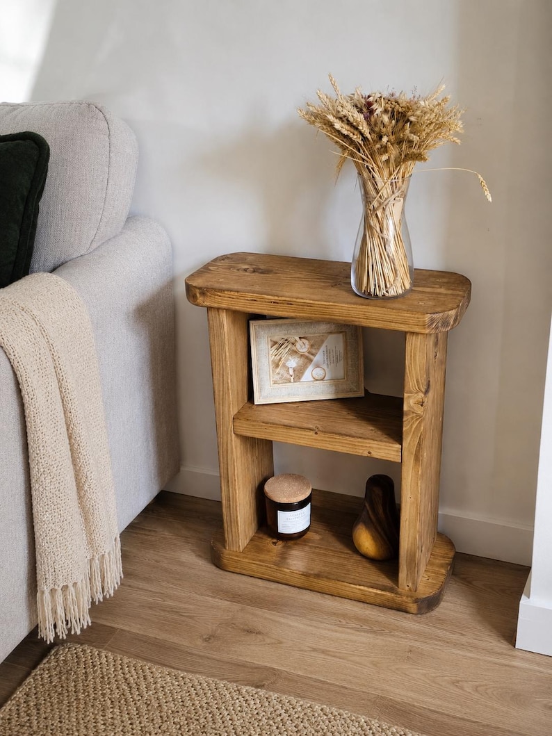 May include: A rustic, two-tiered wooden side table with a warm, honey-toned finish. The table holds a framed picture, a candle, and a decorative wooden pear. A clear glass vase with dried wheat stalks sits on top.
