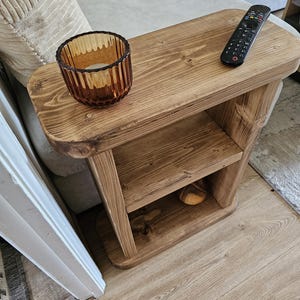 May include: A rustic, wooden side table with a warm, brown finish. It features a top surface with a remote control and a decorative amber glass candle holder. The table has a lower shelf and is designed for placement beside a sofa.