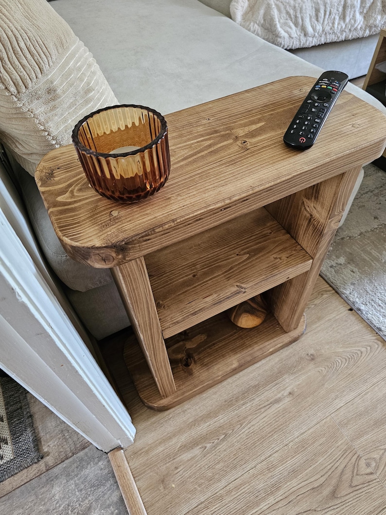 May include: A rustic, wooden side table with a warm, brown finish. It features a top surface with a remote control and a decorative amber glass candle holder. The table has a lower shelf and is designed for placement beside a sofa.