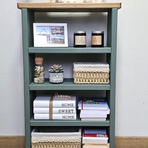 A wooden painted bookshelf with candles, framed photos, two baskets, decorative jars, and stacked books  placed on a wooden floor against a white wall.