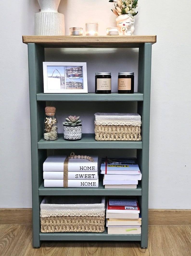 A wooden painted bookshelf with candles, framed photos, two baskets, decorative jars, and stacked books  placed on a wooden floor against a white wall.