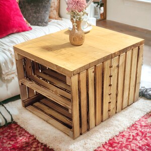 May include: A wooden crate coffee table with a natural wood finish. The table has two shelves and is decorated with a vase of pink flowers.