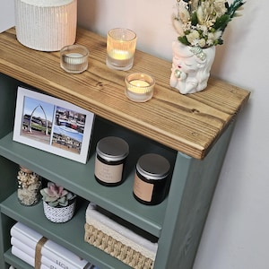 A wooden painted bookshelf with candles, framed photos, two baskets, decorative jars, and stacked books  placed on a wooden floor against a white wall.