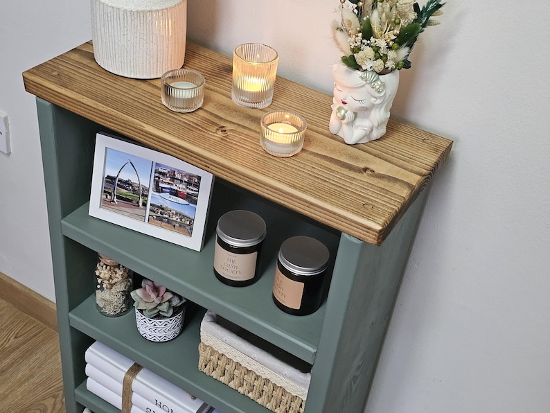 A wooden painted bookshelf with candles, framed photos, two baskets, decorative jars, and stacked books  placed on a wooden floor against a white wall.