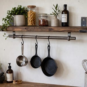May include: A dark brown wooden wall shelf with a metal rail and three black S-hooks holding one stainless steel saucepan and two black cast iron skillets. The shelf holds a potted plant, two glass jars with pasta and spices, and a bottle.