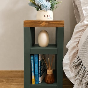 May include: A dark green bedside table with a wooden top. It features two shelves, one holding books and a diffuser, the other a vase. A vase of blue and white flowers sits on top. The word "Life" is written on the vase.