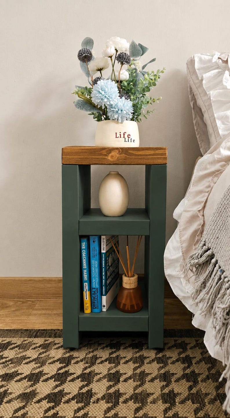 May include: A dark green bedside table with a wooden top. It features two shelves, one holding books and a diffuser, the other a vase. A vase of blue and white flowers sits on top. The word "Life" is written on the vase.