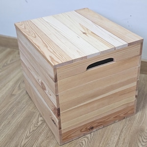 May include: A light-coloured wooden storage box with a rectangular shape. The box is constructed from horizontal wooden planks and features a handle cut into one side. The box is sitting on a wooden floor.