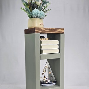 May include: A sage green wooden side table with two shelves. The top shelf holds a cream-colored pot with the word "Life" and a floral arrangement. The middle shelf has three books, and the bottom shelf displays a decorative sailboat.