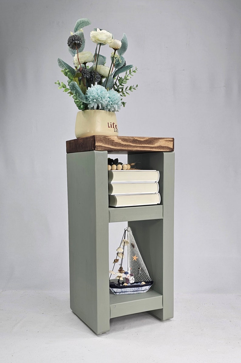 May include: A sage green wooden side table with two shelves. The top shelf holds a cream-colored pot with the word "Life" and a floral arrangement. The middle shelf has three books, and the bottom shelf displays a decorative sailboat.