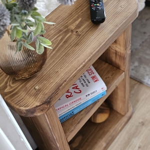 May include: A small, rustic wooden side table with two shelves. The table is stained a warm brown and has rounded edges. A black remote control sits on top, and two books are visible on the middle shelf. A vase with flowers is on the top shelf.